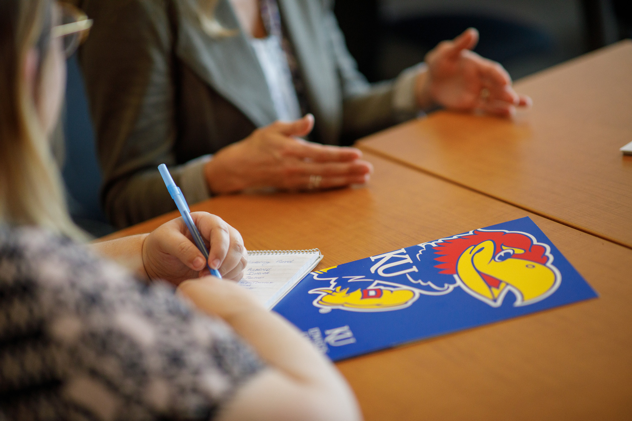 a seated individual is writing on a notepad at a table with another individual in the background 